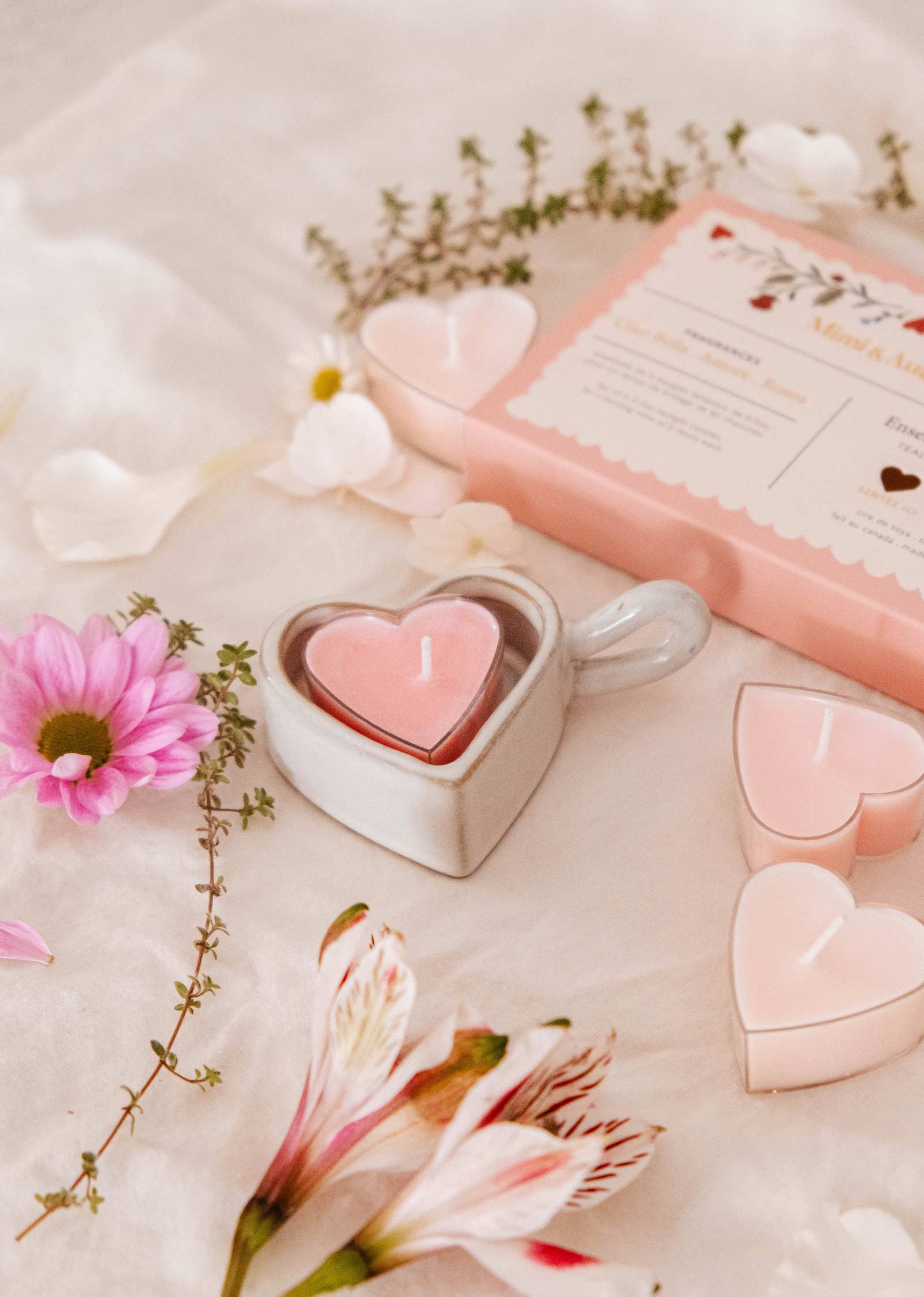 Le porte-bougies à réchaud Heart & Glow de Mimi & August et une boîte à fleurs rose reposent sur un tissu, entourés de bougies à réchaud blush hearts, de marguerites roses, de fleurs blanches et de verdure luxuriante pour un décor romantique parfait.