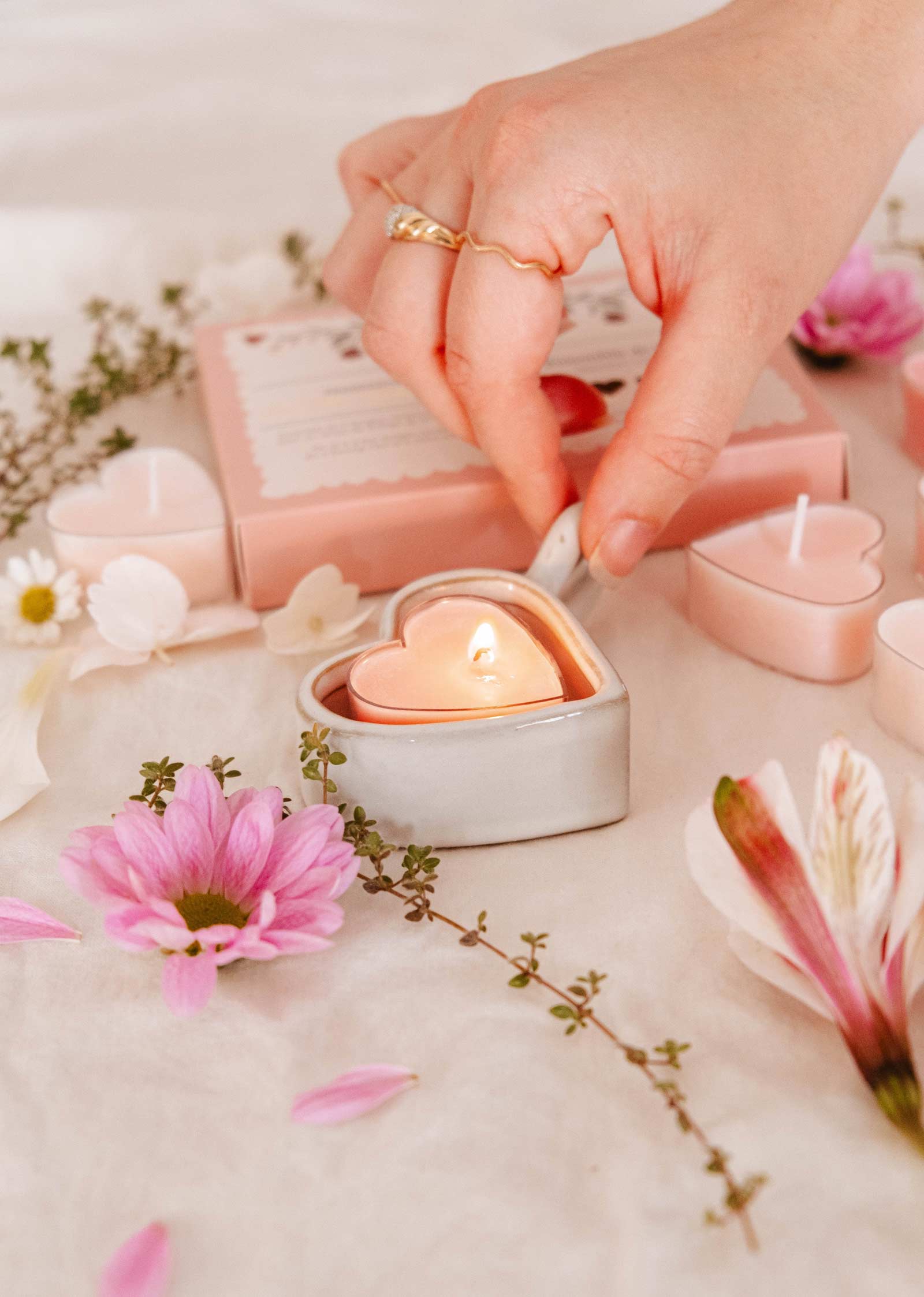 A hand lights a candle, part of a romantic decor, surrounded by flowers and unlit Heart & Glow Tealight Holders from Mimi & August on a white surface.