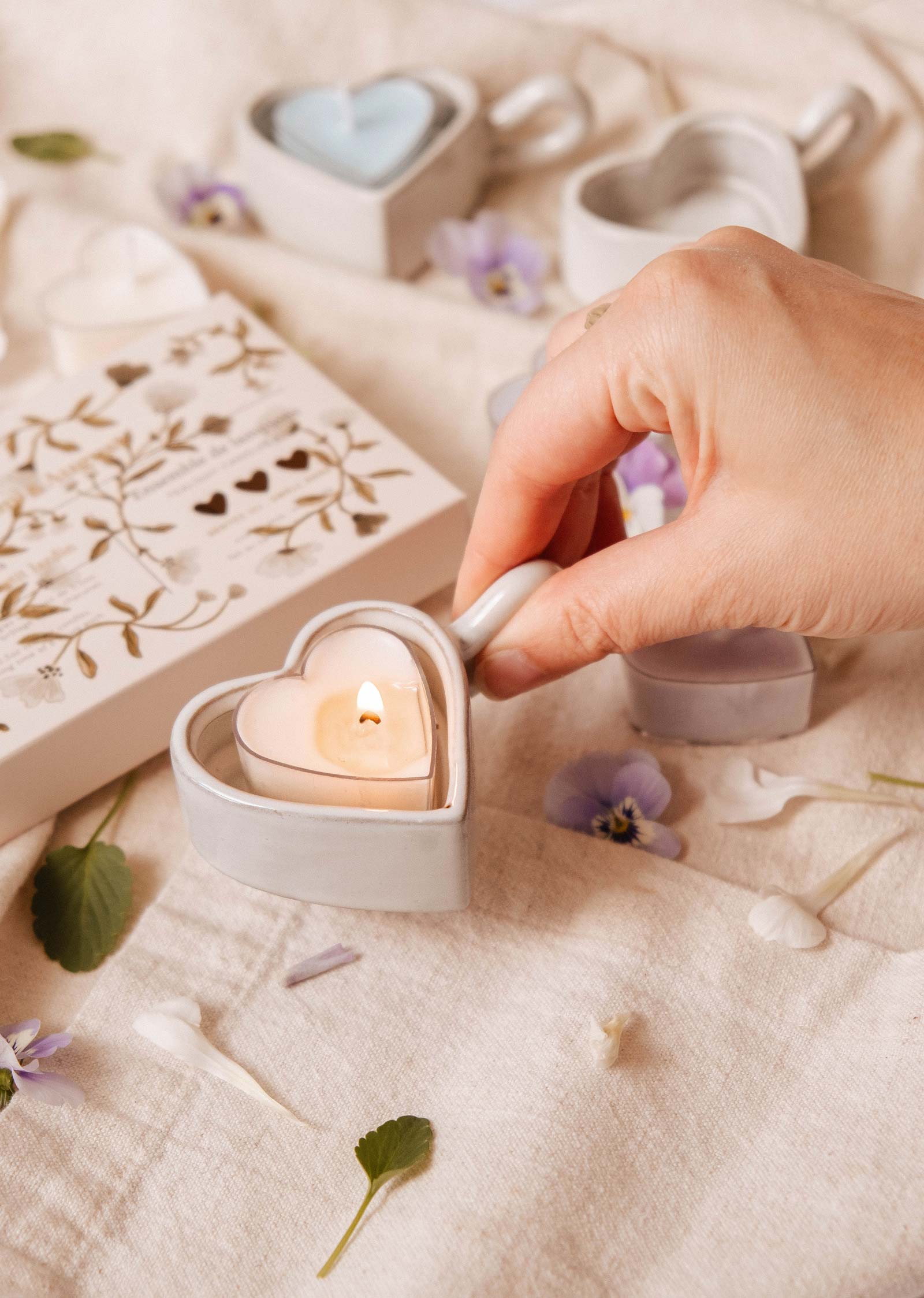 Une main tient le porte-bougies à réchaud Mimi & August Heart & Glow avec une bougie allumée à l'intérieur, entourée de bougies à réchaud Blush Hearts, de fleurs et d'une boîte décorative sur une surface en tissu léger.