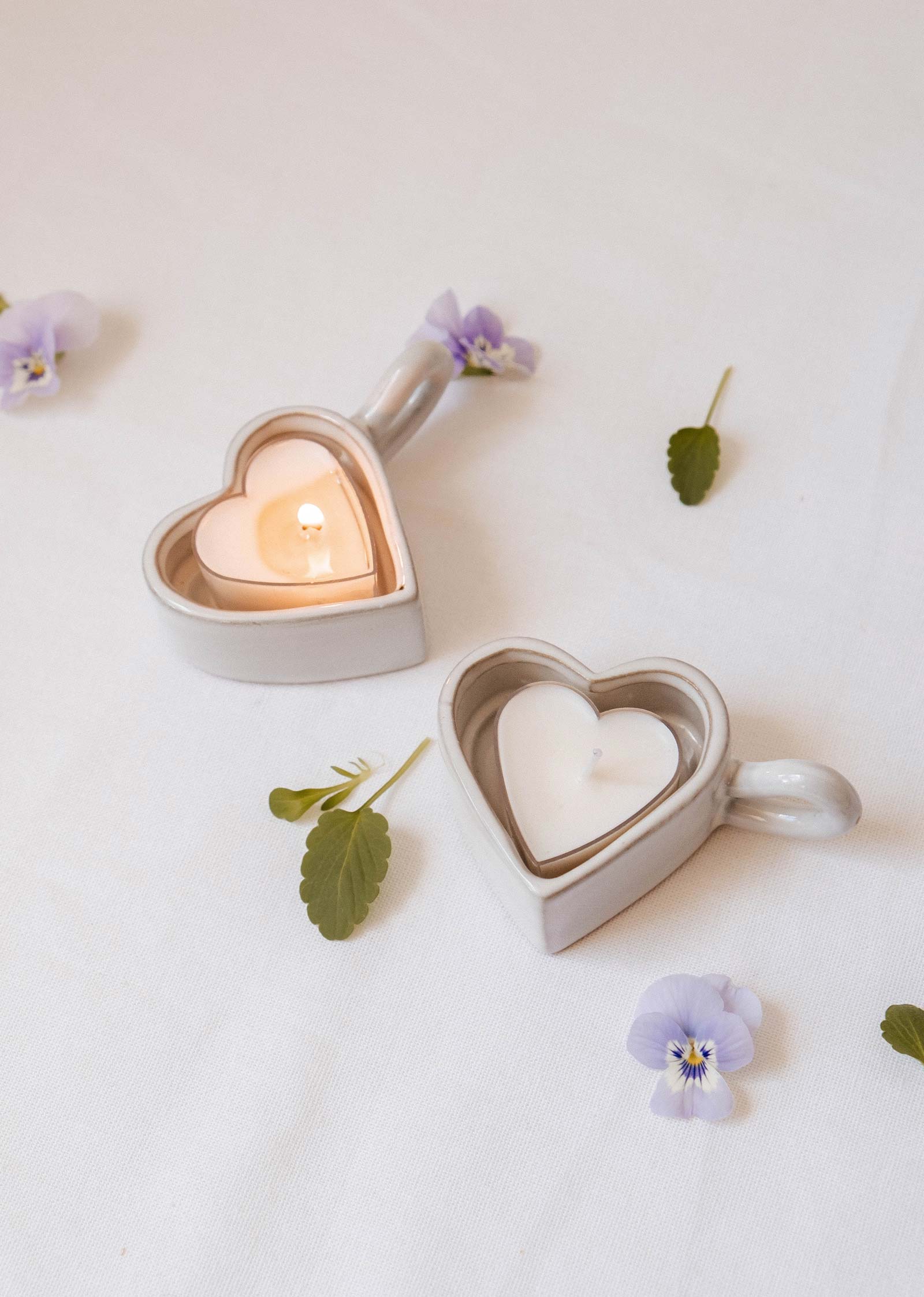 Two Heart & Glow Tealight Holders by Mimi & August, one with a lit and one with an unlit tealight, sit on a white cloth decorated with scattered purple flowers and green leaves.