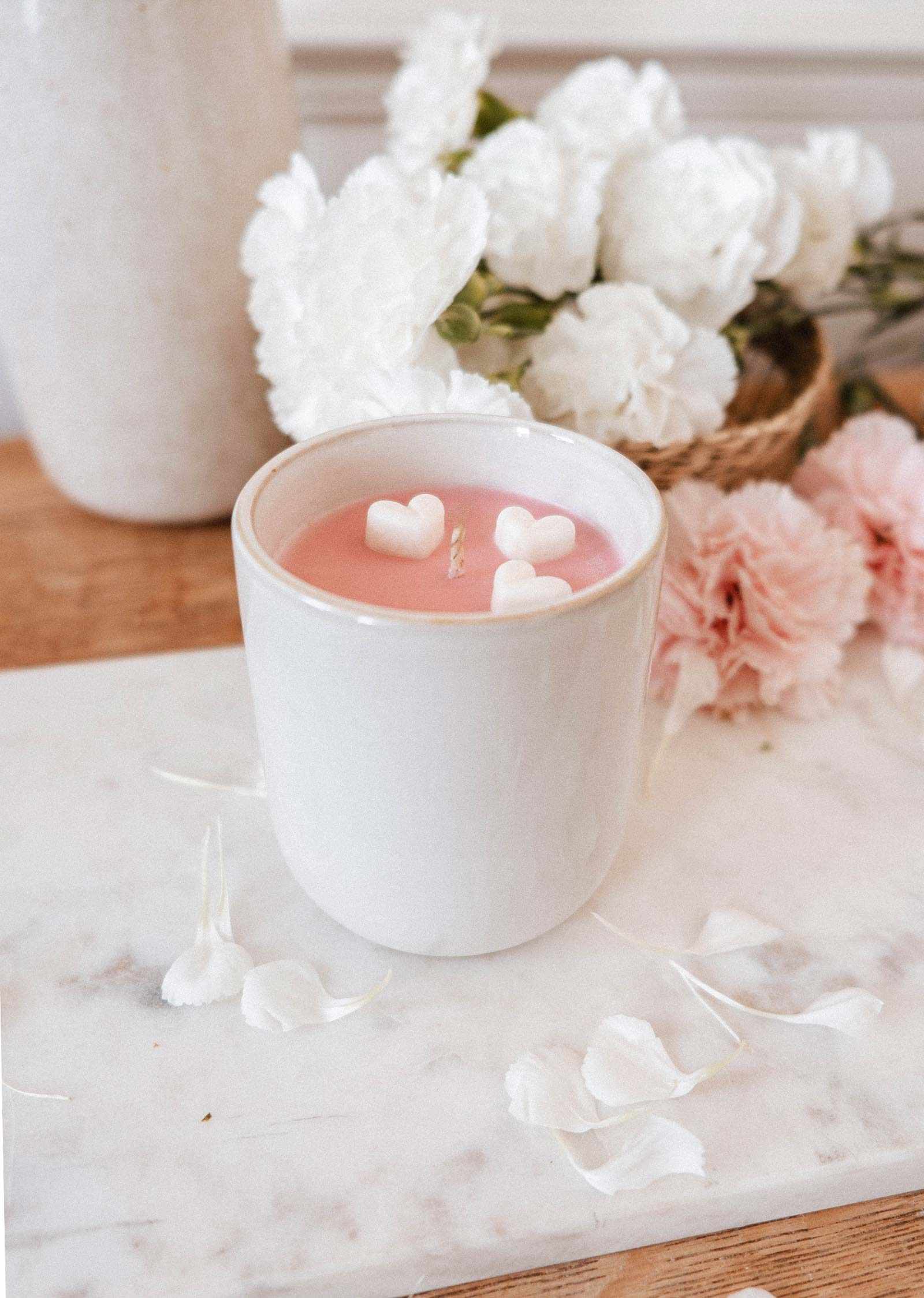 The Mimi & August Special Edition Candle features pink wax with heart-shaped pieces in a white cup, placed on marble and surrounded by white and pink flowers with scattered petals.