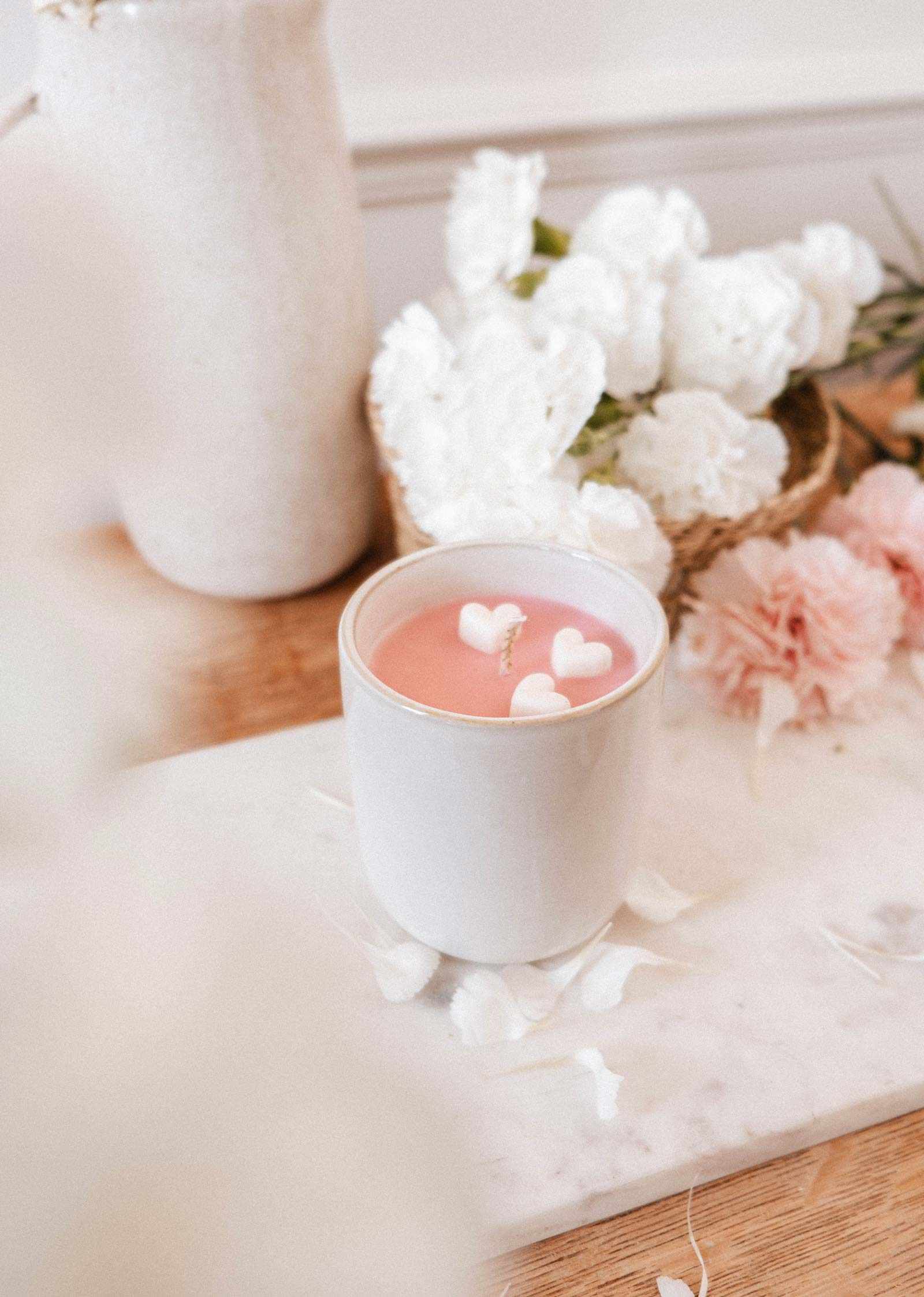 The Special Edition Candle by Mimi & August, a pink limited edition candle in a white cup with heart-shaped wax pieces, sits on marble among white and pink flowers.