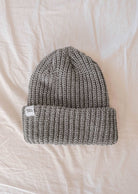 Le bonnet Heather Grey Chunky Beanie de Mimi & August, méticuleusement confectionné avec un fil épais, présente un bord plié et une petite étiquette blanche, le tout posé élégamment sur une surface blanche - un accessoire d'hiver parfait.