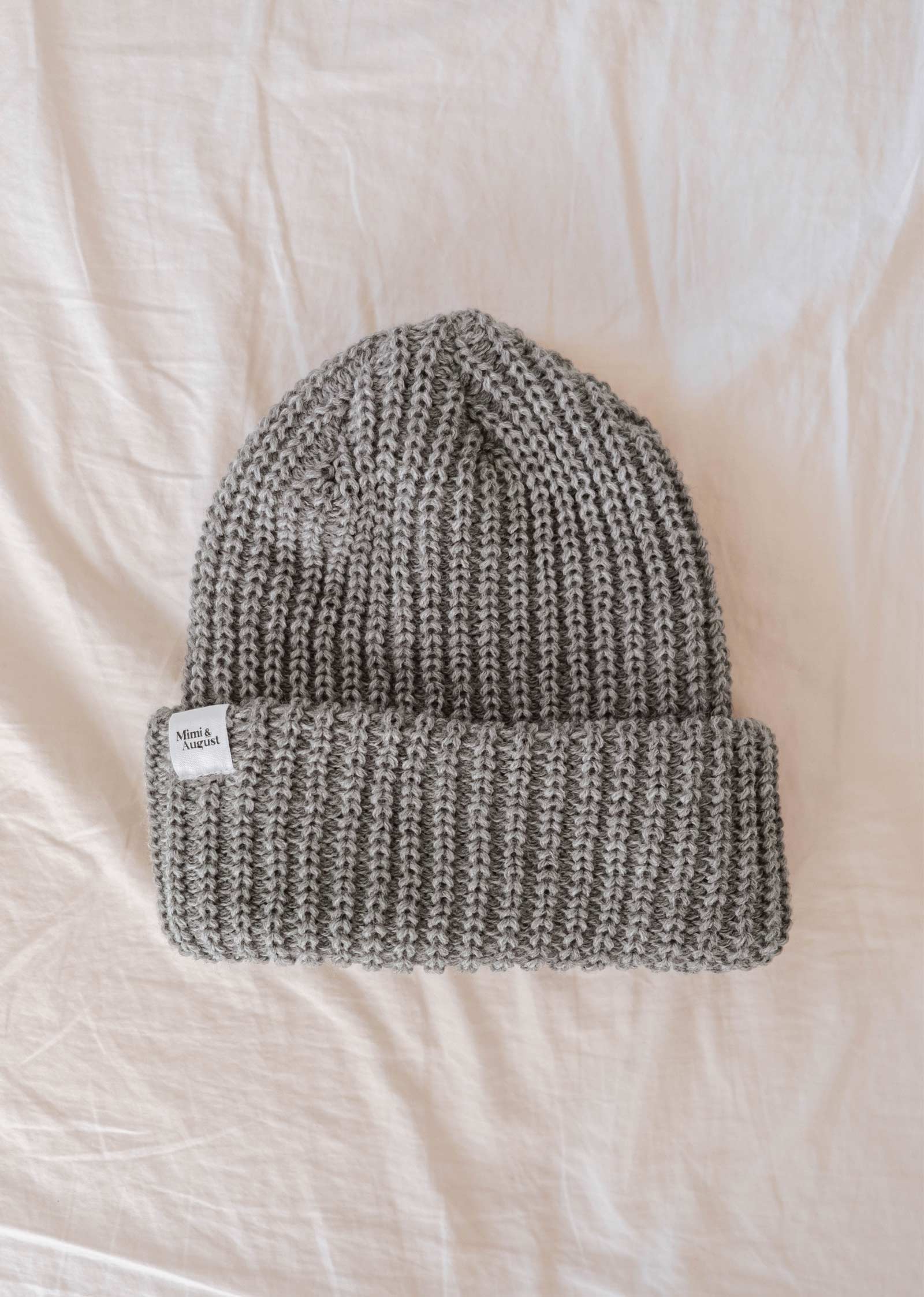 Le bonnet Heather Grey Chunky Beanie de Mimi & August, méticuleusement confectionné avec un fil épais, présente un bord plié et une petite étiquette blanche, le tout posé élégamment sur une surface blanche - un accessoire d'hiver parfait.