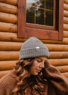 Une personne aux cheveux longs et ondulés, portant un bonnet Heather Grey Chunky Beanie de Mimi & August et un pull marron, se tient devant le mur et la fenêtre d'une cabane en bois.