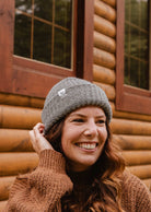 Une femme portant un bonnet Mimi & August Heather Grey Chunky Beanie et un pull marron sourit devant une cabane en bois.