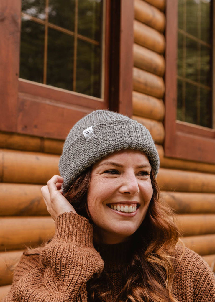 A woman wearing a Mimi & August Heather Grey Chunky Beanie and a brown sweater smiles in front of a wooden cabin.