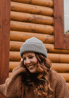 Une personne portant un bonnet Heather Grey Chunky Beanie de Mimi & August et un pull se tient devant une cabane en rondins avec un sourire.