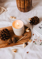 Une bougie allumée **Heirloom - Reusable Candle** de **Mimi & August** dans un support en céramique est posée sur un plateau en bois avec des pommes de pin, un marron, une allumette et un échantillon de parfum. Le plateau est posé sur un tissu blanc à côté d'un livre.