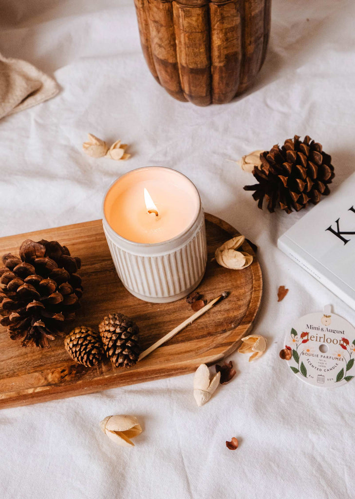 A lit **Heirloom - Reusable Candle** by **Mimi & August** in a ceramic holder sits on a wooden tray with pinecones, chestnut, a matchstick, and fragrance sample. The tray is placed on a white cloth next to a book.