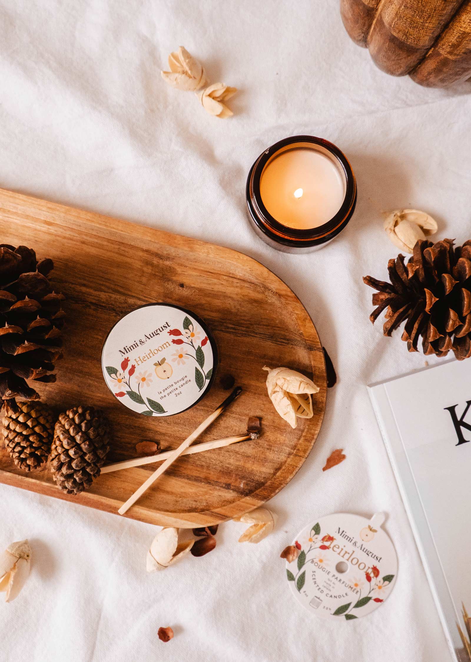 Un plateau en bois orné de pommes de pin, la bougie réutilisable Heirloom de Mimi & August dégageant un riche parfum de cèdre, et de petits objets décoratifs sur une surface blanche, à côté d'un magazine ou d'un livre partiellement visible.