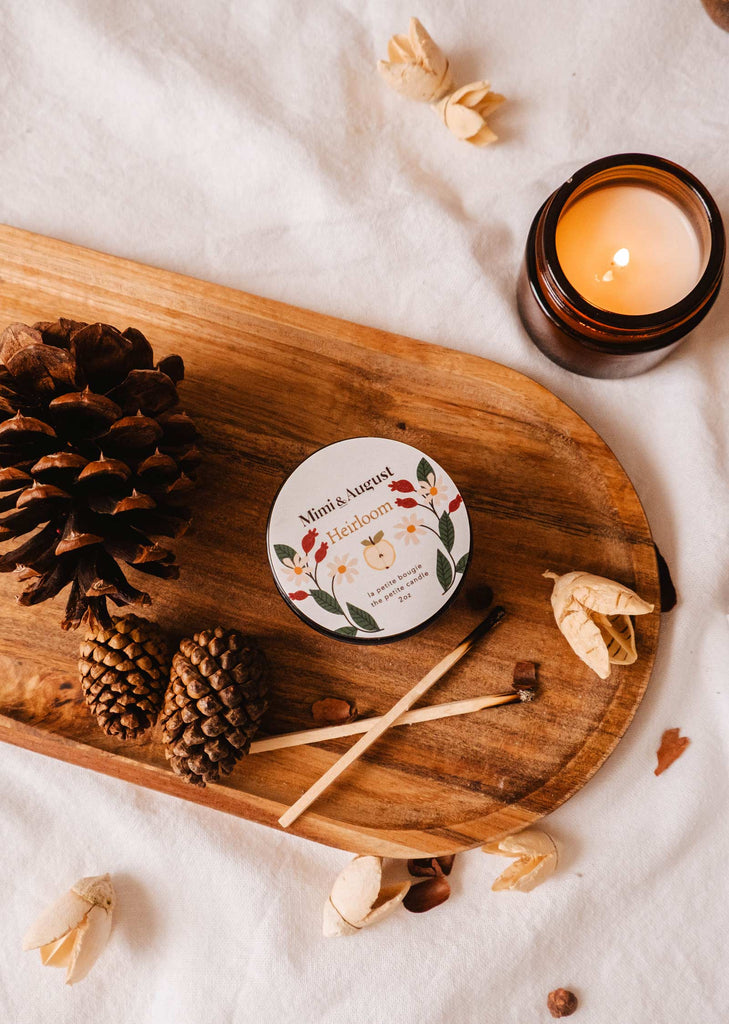 A lit Heirloom - Reusable Candle by Mimi & August, a container labeled "Heirloom," pinecones, and matches are arranged on a wooden tray placed on a white fabric background, with the subtle scents of cedar and clove enhancing the cozy ambiance.