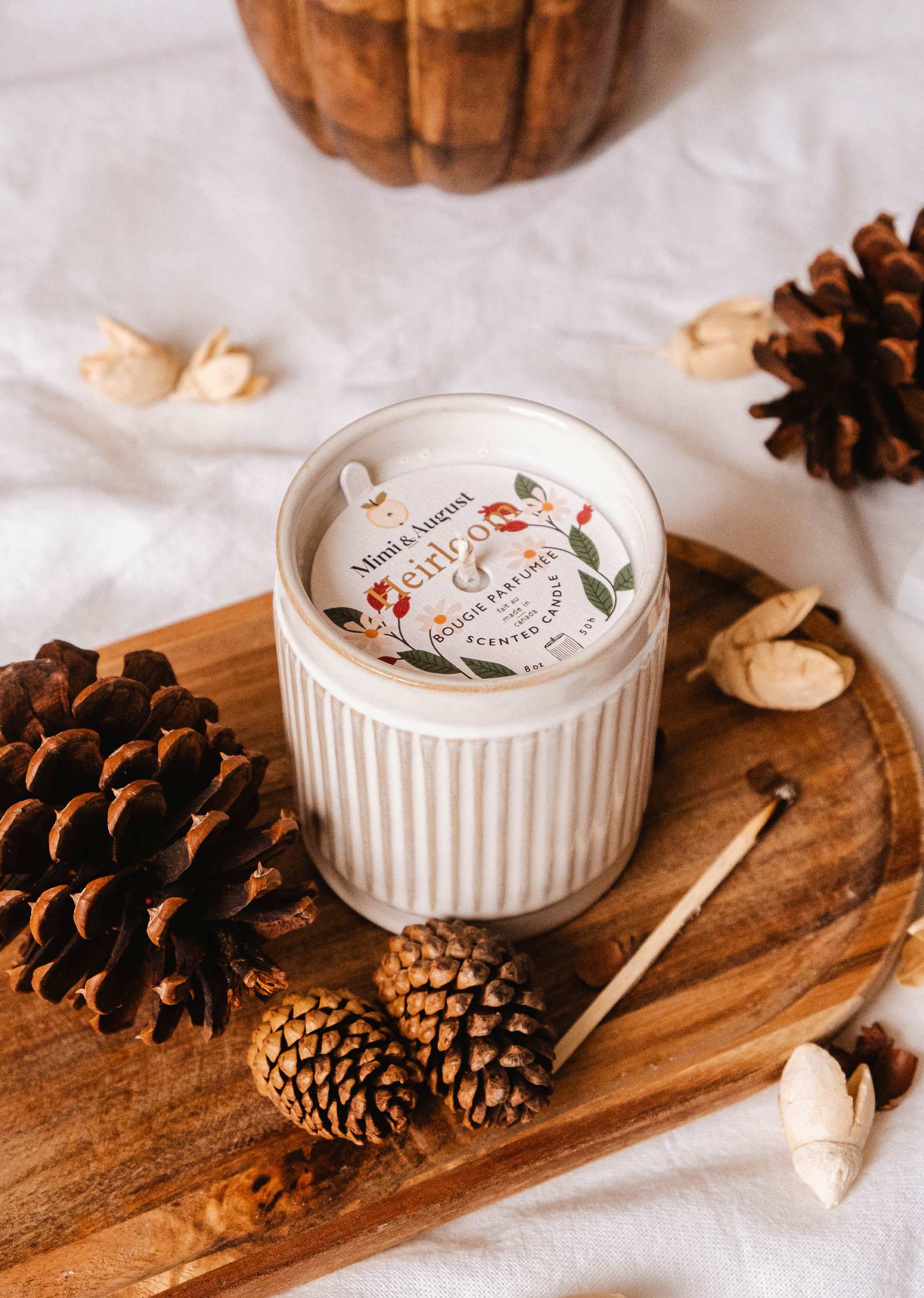 Une bougie réutilisable Heirloom de Mimi & August, logée dans un support en céramique blanche, est posée sur un plateau en bois entouré de pommes de pin, d'accents de châtaigne et de touches de cèdre. Un bâton d'allumette est posé à côté de la bougie.