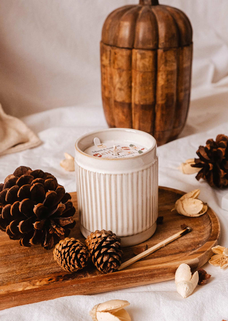 A white ribbed Heirloom Reusable Candle from Mimi & August sits on a wooden tray with pinecones, cedar matches, and dried petals, accompanied by a wooden pumpkin ornament in the background.