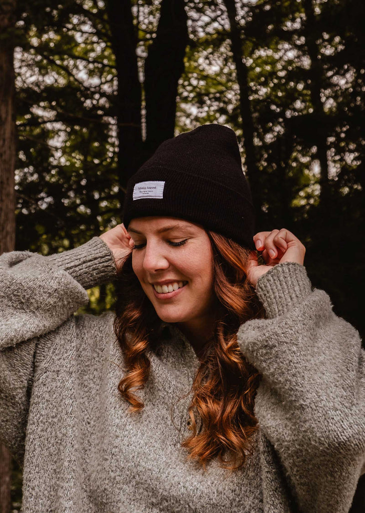 A person wearing a gray sweater and an Intemporel Black Beanie from Mimi & August smiles while standing in a forested area, showcasing their chic winter accessory.