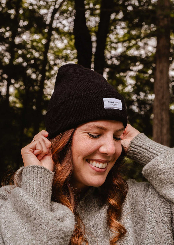 A person with long curly hair, wearing a Mimi & August Intemporel Black Beanie and a knitted sweater, smiles while standing outdoors with trees in the background.