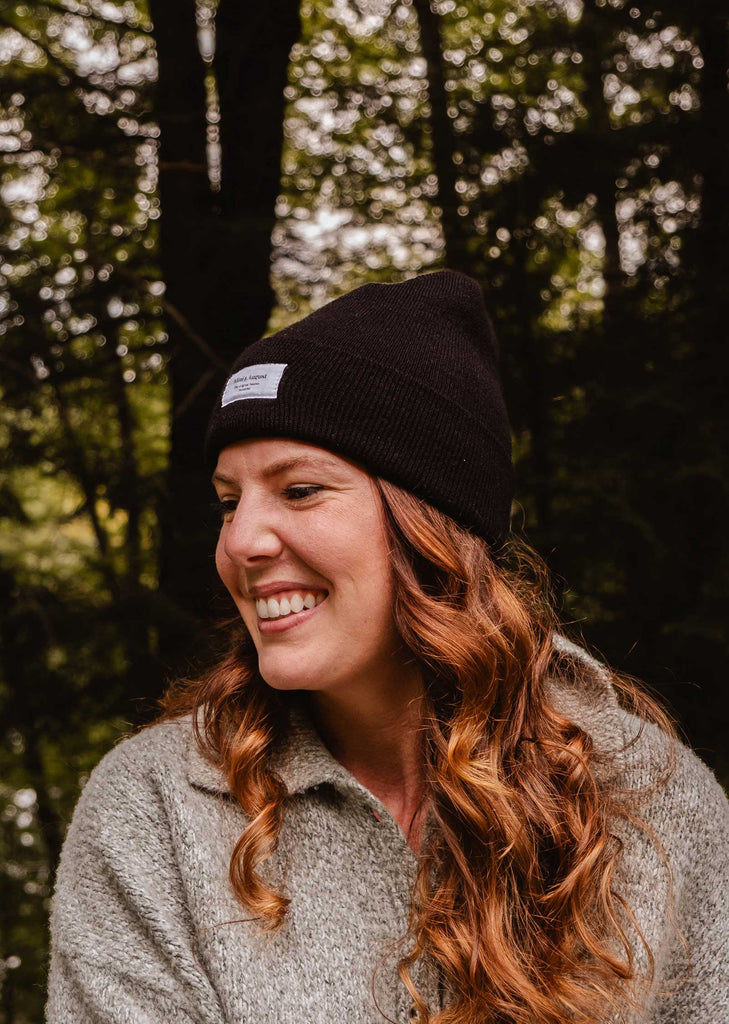 A woman with long, wavy hair wearing a Mimi & August Intemporel Black Beanie and a grey sweater smiles while outdoors. Trees are visible in the background, highlighting her stylish winter accessory.