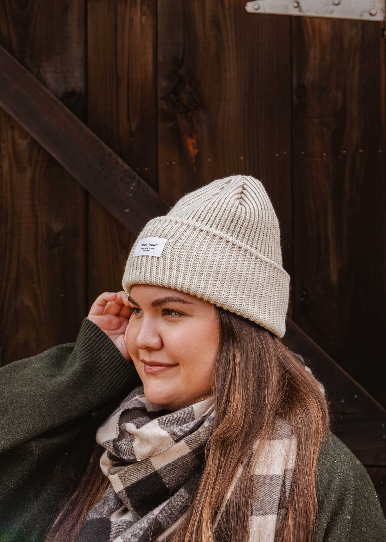 Une personne portant un bonnet en tricot de couleur claire, un pull vert et une écharpe à carreaux se tient devant un fond en bois, mettant en valeur sa garde-robe d'hiver avec le bonnet unisexe Ivory Snuggle Beanie de Mimi & August.