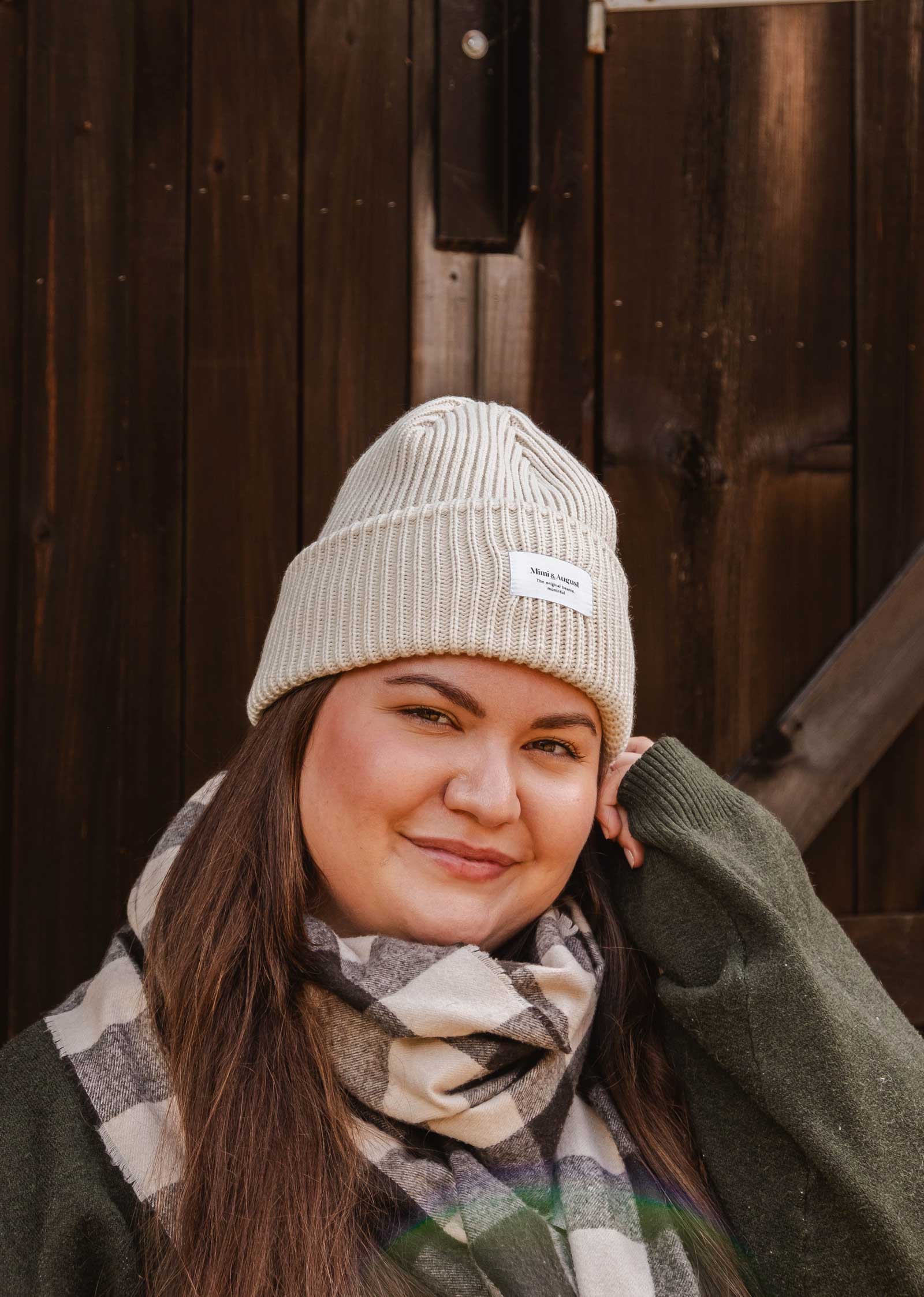 Une personne portant un bonnet Ivory Snuggle Beanie de Mimi & August, un pull vert et une écharpe à carreaux pose devant un fond en bois, mettant en valeur leur tricot acrylique unisexe pour parfaire leur garde-robe hivernale.