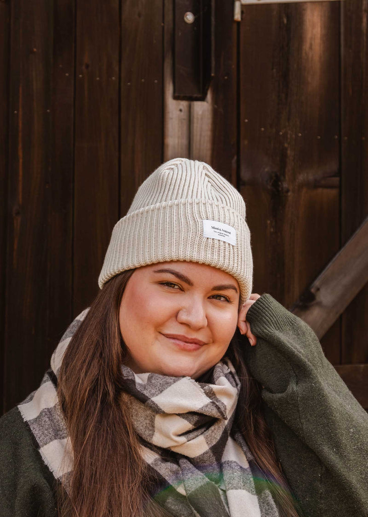 A person wearing a Mimi & August Ivory Snuggle Beanie, a green sweater, and a checkered scarf poses in front of a wooden background, showcasing their unisex acrylic knit to perfect their winter wardrobe.