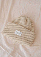 The Ivory Snuggle Beanie by Mimi & August, a beige ribbed accessory featuring a white logo patch, rests on a cream-colored fabric surface, making it an ideal and versatile addition to any winter wardrobe. This unisex acrylic knit ensures both style and warmth.