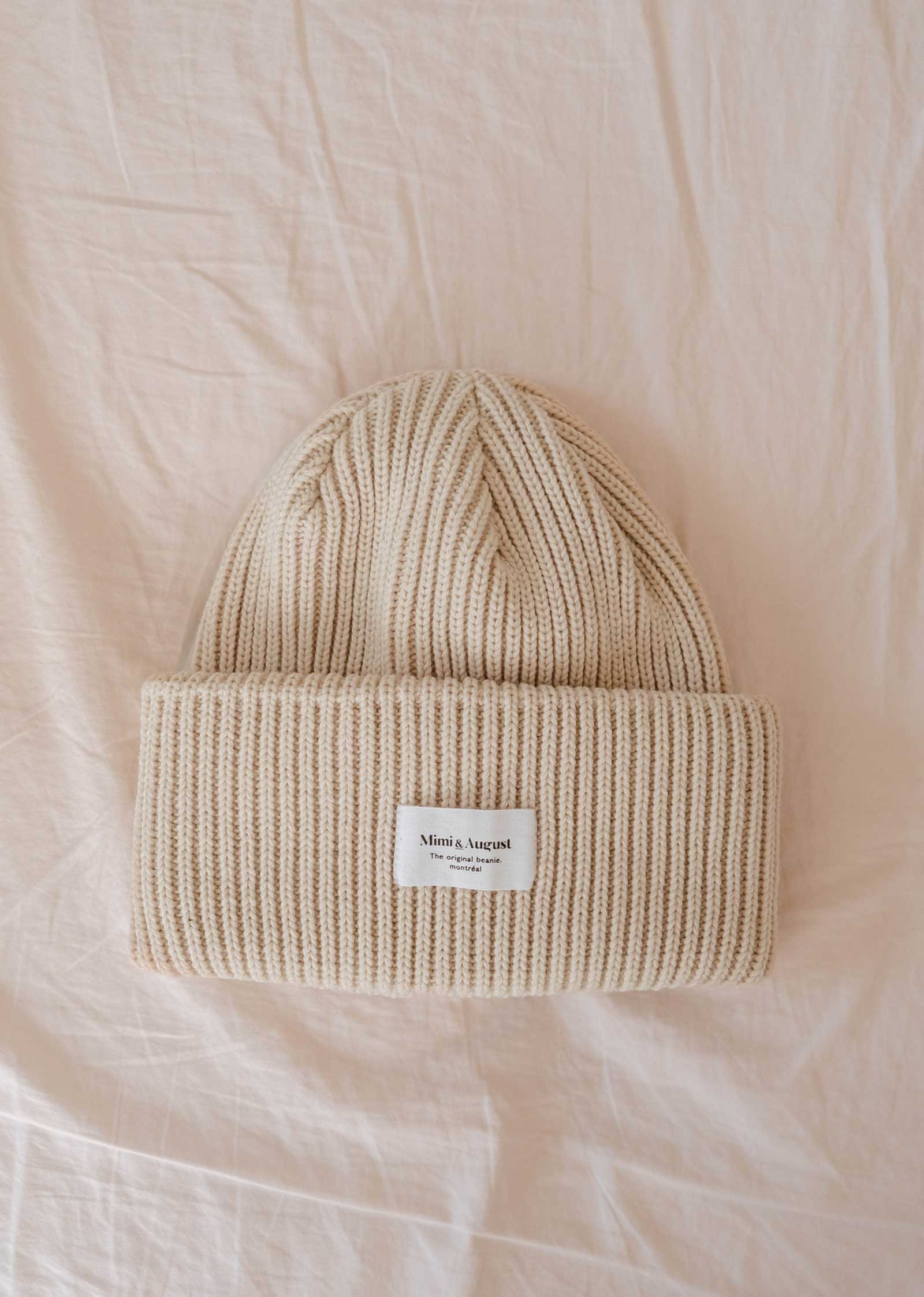 Le bonnet Ivory Snuggle Beanie est un bonnet beige en maille côtelée portant une étiquette blanche avec "Mimi & August" sur un fond de tissu crème. Ce bonnet unisexe en acrylique est indispensable à votre garde-robe d'hiver.