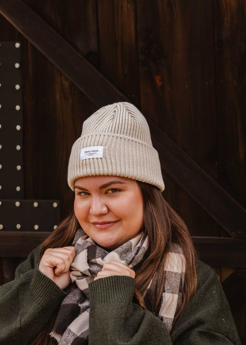 A person wearing a Mimi & August Ivory Snuggle Beanie and a checkered scarf stands in front of a dark wooden background, looking at the camera, showcasing their winter wardrobe.