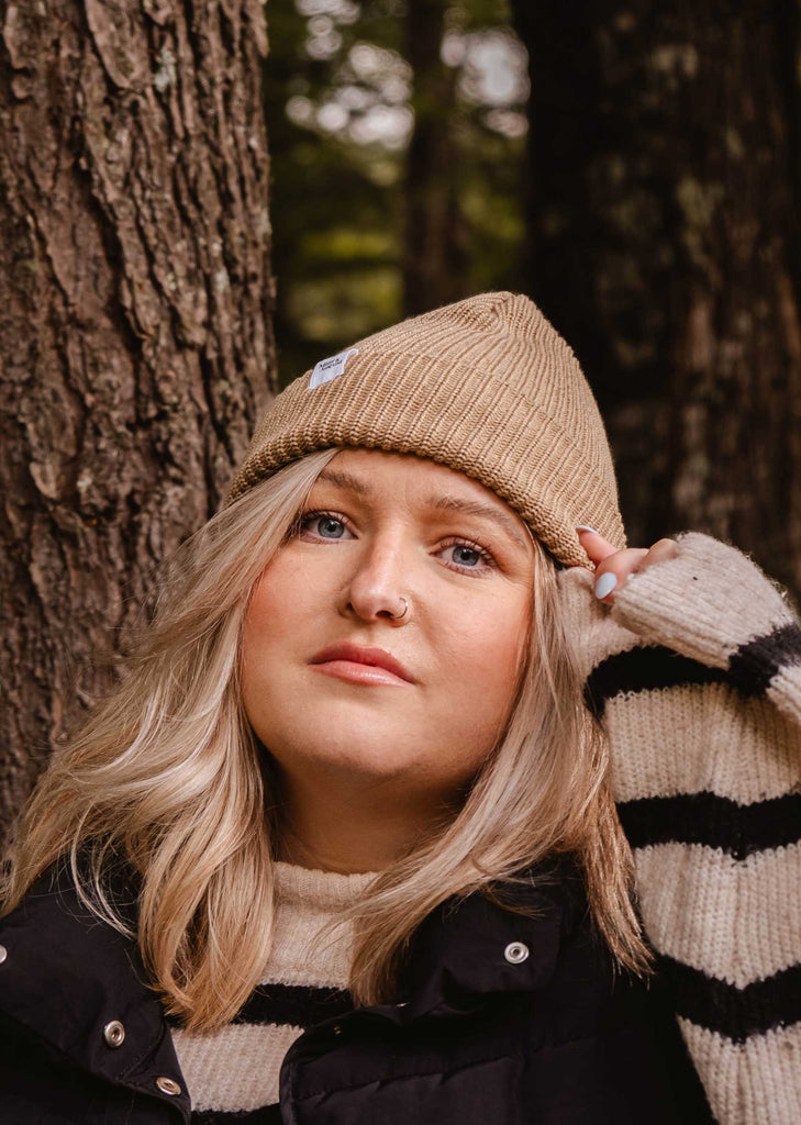 A person is standing next to a tree, wearing a black jacket over a striped sweater and topped with the Khaki Cap Super Puff Beanie by Mimi & August, making for the perfect winter accessory.