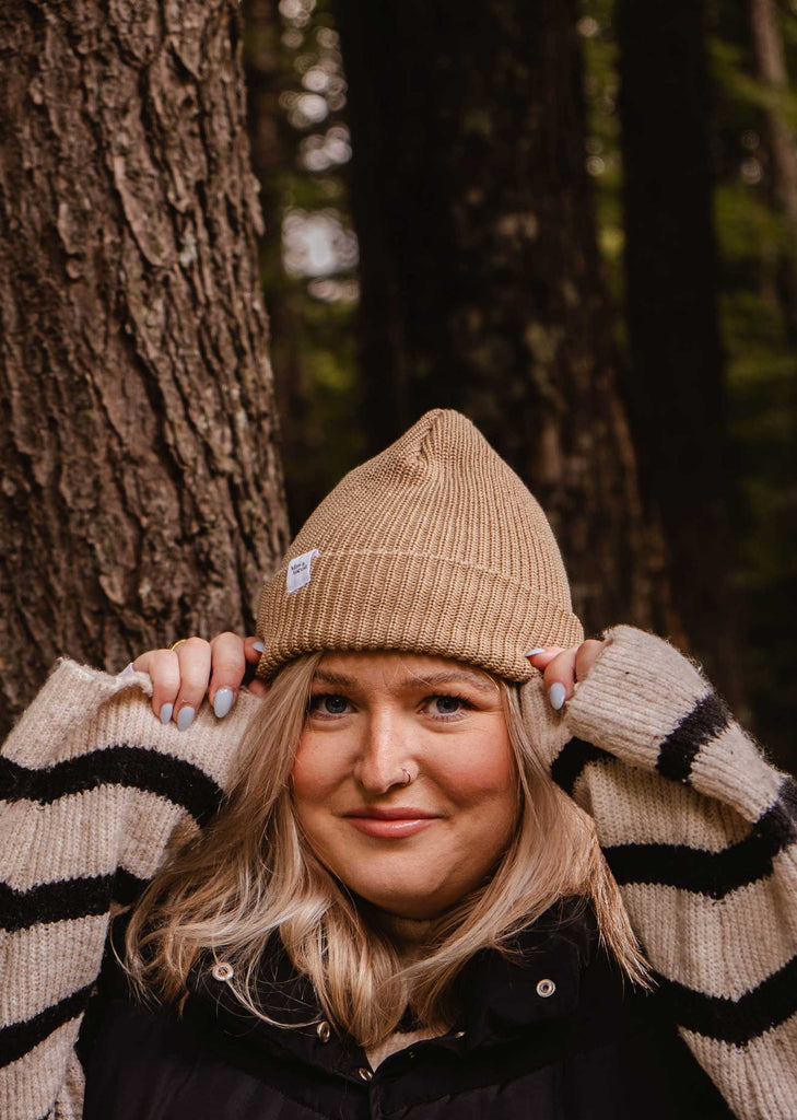 A person wearing a Mimi & August Khaki Cap Super Puff Beanie and a striped sweater stands in front of a tree in a forest, showcasing the perfect winter accessory.