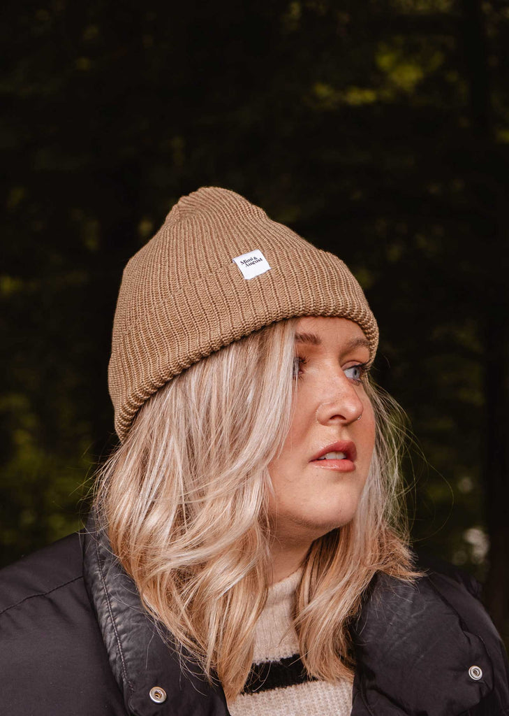 A person with blonde hair wearing a Mimi & August Khaki Cap Super Puff Beanie and a black jacket, perfect as a winter accessory, is glancing to the side against a blurred outdoor background.