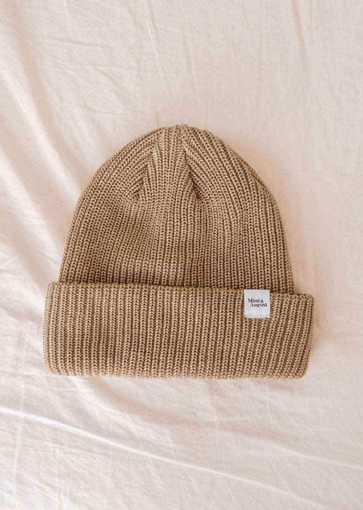 A folded light brown knitted beanie hat, the Khaki Cap Super Puff Beanie by Mimi & August, with a small white tag on the brim, rests on a cream-colored fabric background. This unisex knit winter accessory seamlessly blends style and warmth.