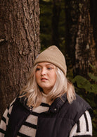 Une personne aux longs cheveux blonds portant un bonnet Mimi & August Khaki Cap Super Puff Beanie, un gilet noir et un pull rayé se tient devant un arbre dans une forêt.