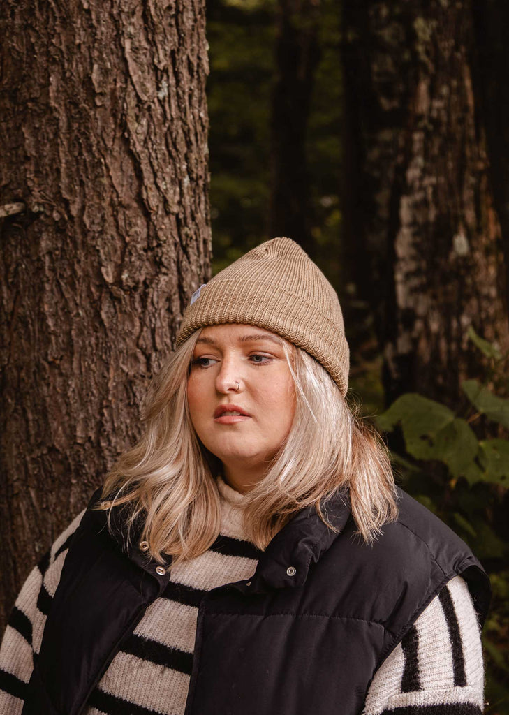 A person with long, blonde hair wearing a Mimi & August Khaki Cap Super Puff Beanie, a black puffer vest, and a striped sweater stands in front of a tree in a forest setting.