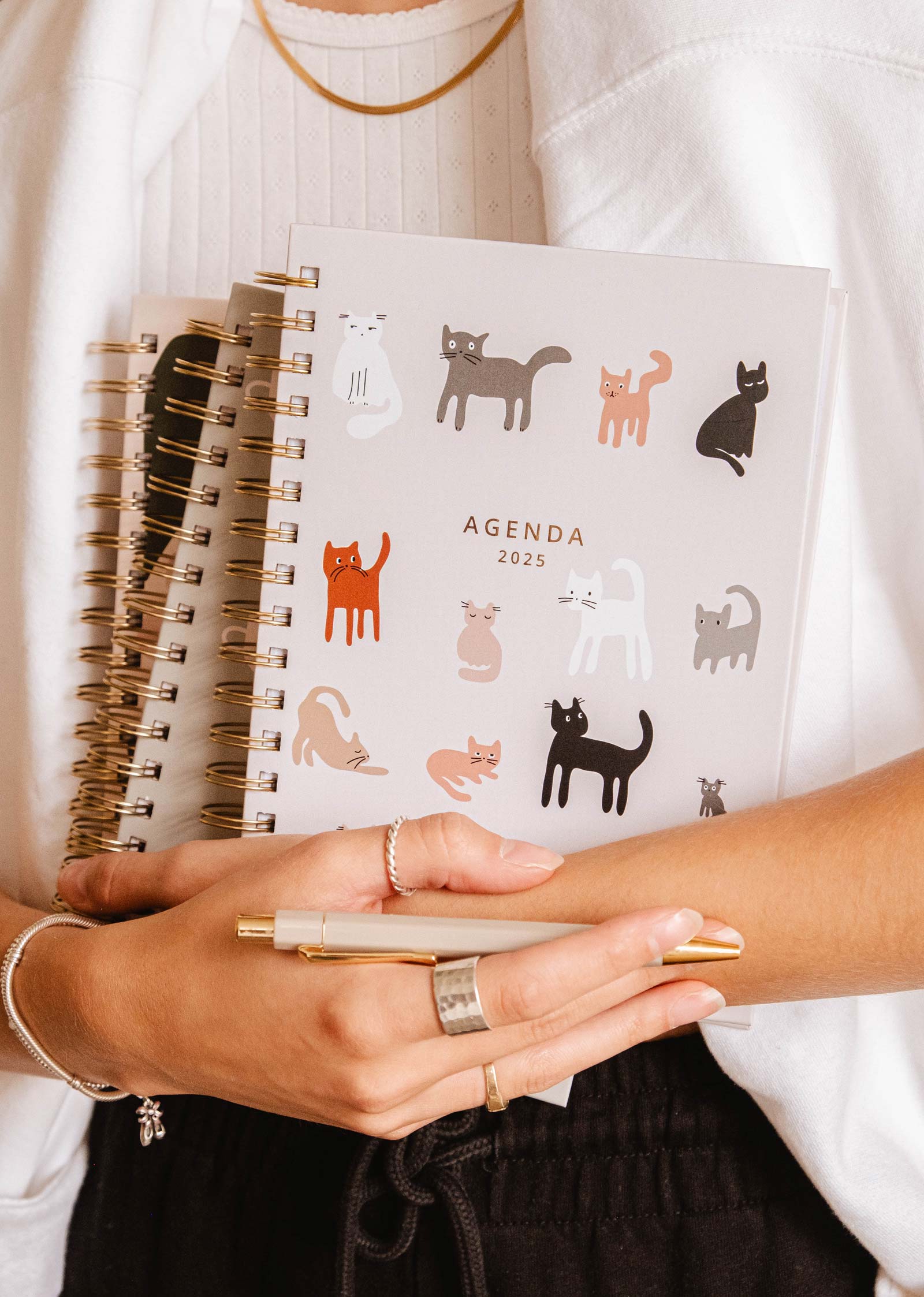 A person holds a 2025 Kittens Agenda by Mimi & August, featuring hand-drawn cat and floral illustrations on the cover, while a pen and another spiral-bound notebook lie beside them to help stay productive.