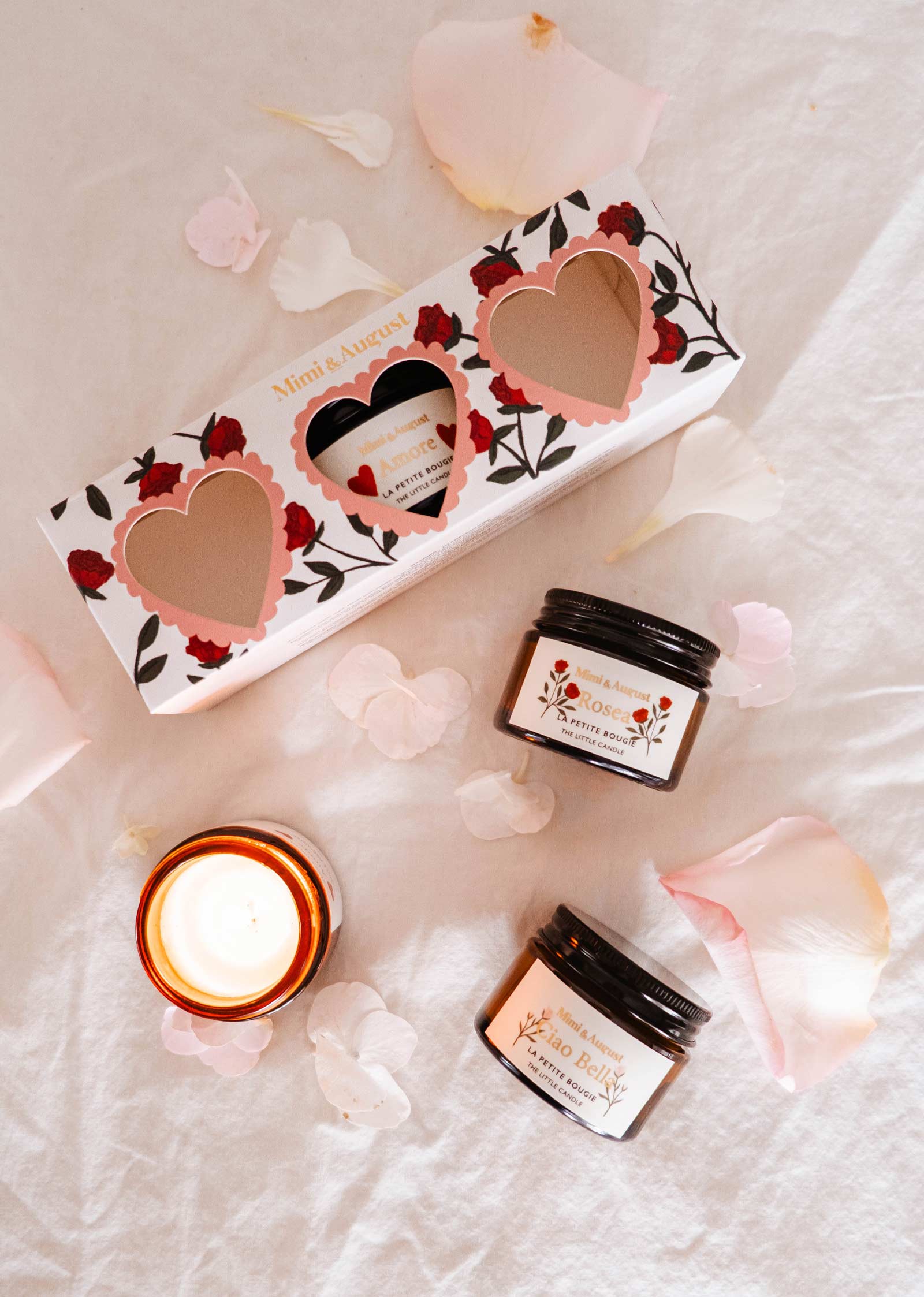 Des articles de soin, dont deux pots et une élégante bougie, reposent sur un tissu rose doux parsemé de pétales, à côté du coffret Mimi & August La Romance, orné de découpes en forme de cœur. Avec des parfums exclusifs, cette collection ajoute du luxe à votre routine de beauté.