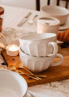 Two stacked Large Ceramic Latte Mug by Mimi & August sit on a wooden board beside gold spoons, a lit tealight candle, and other tableware on a white tablecloth.