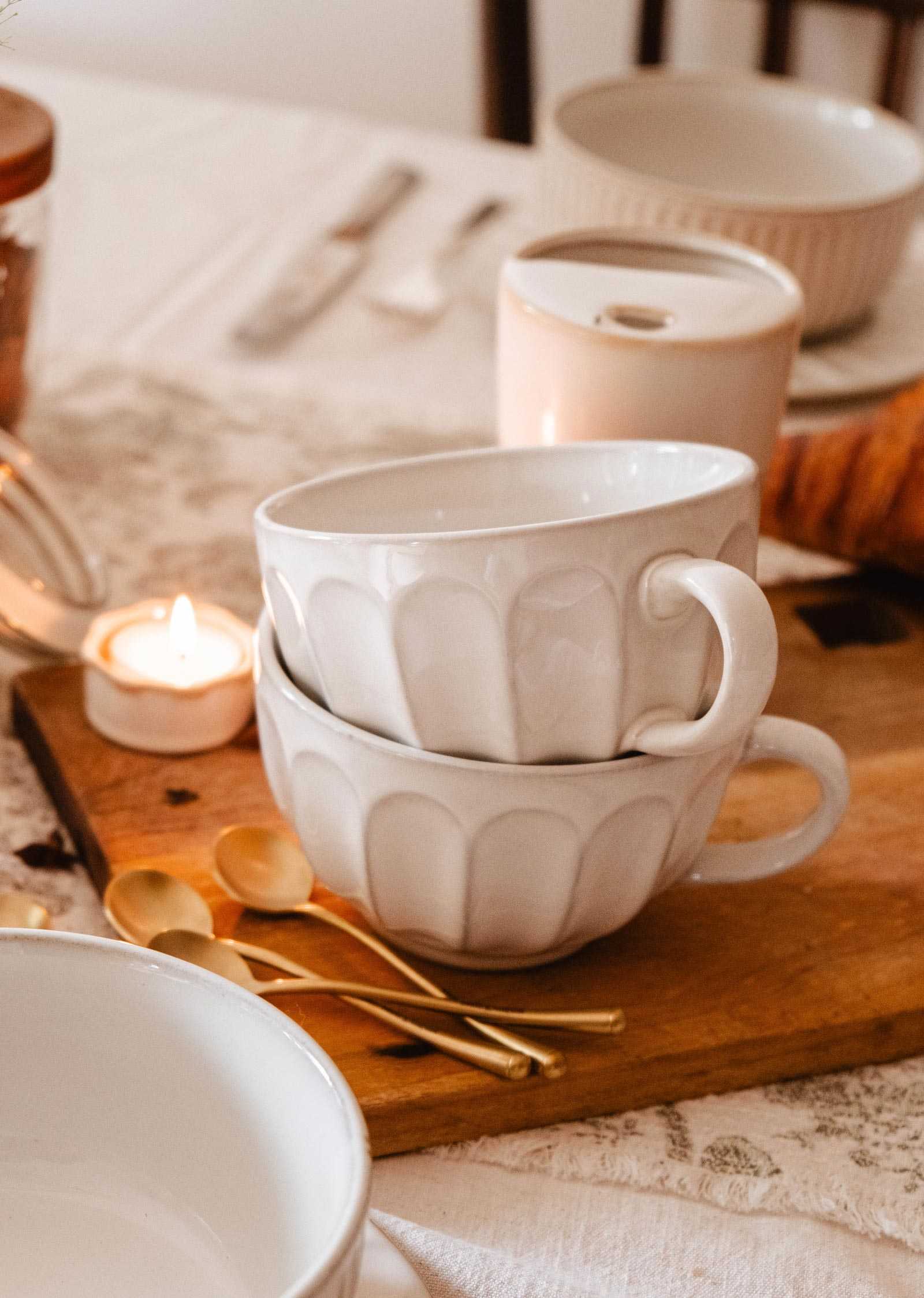 Two stacked Large Ceramic Latte Mug by Mimi & August sit on a wooden board beside gold spoons, a lit tealight candle, and other tableware on a white tablecloth.