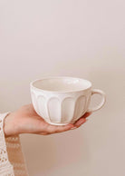 A hand holds a Ceramic Latte Mug by Mimi & August, displaying its white ceramic body with a textured, scalloped pattern against a plain light background.