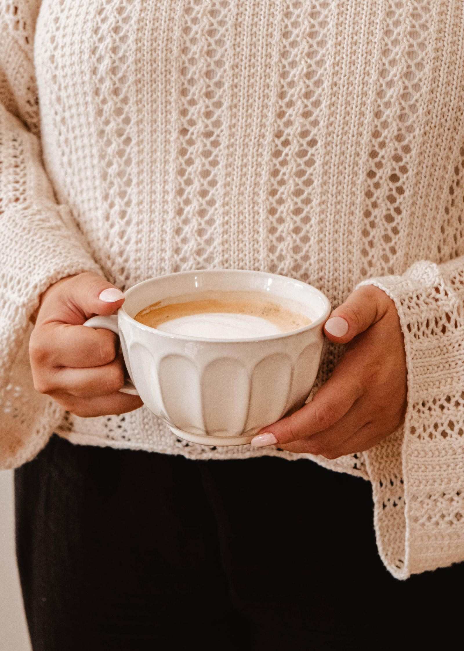 A person wearing a cream knit sweater holds the Large Ceramic Latte Mug by Mimi & August, filled with a latte or cappuccino, with both hands visible.