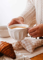 A person in a knit sweater holds The Ceramic Latte Mug by Mimi & August, filled with coffee, above a table set with a croissant, a checked napkin, and a white bowl.