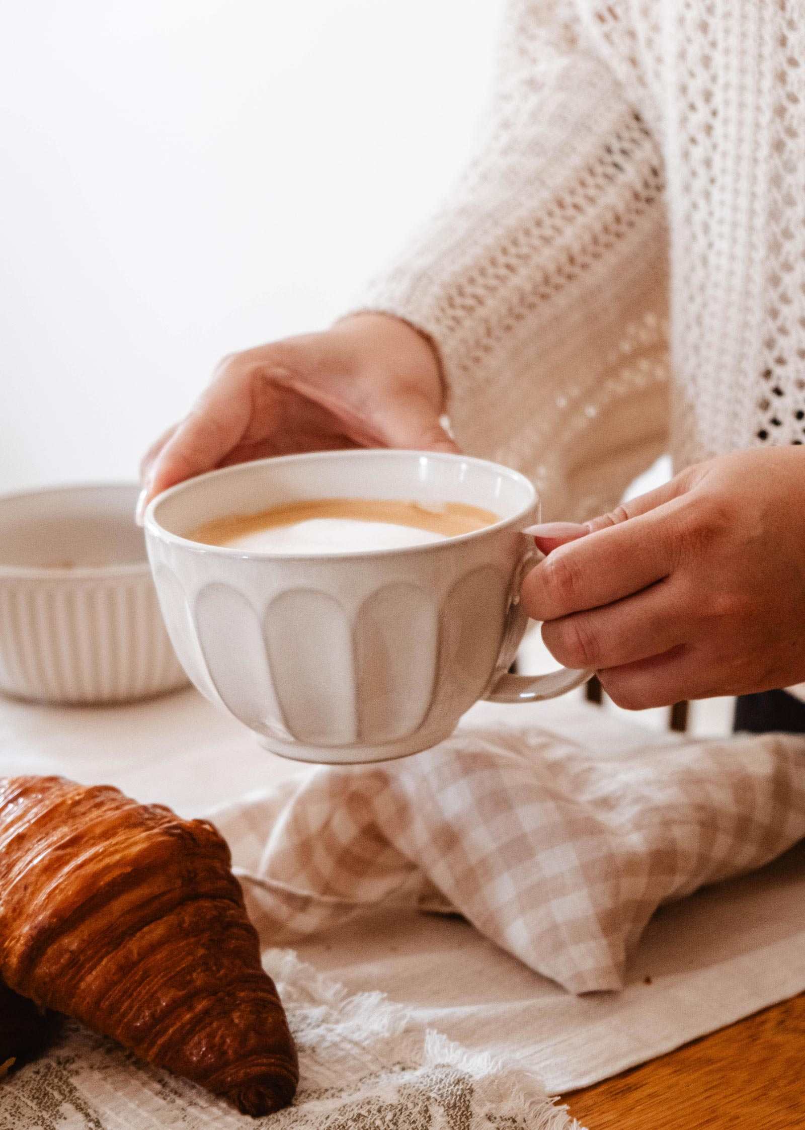 A person in a knit sweater holds The Ceramic Latte Mug by Mimi & August, filled with coffee, above a table set with a croissant, a checked napkin, and a white bowl.