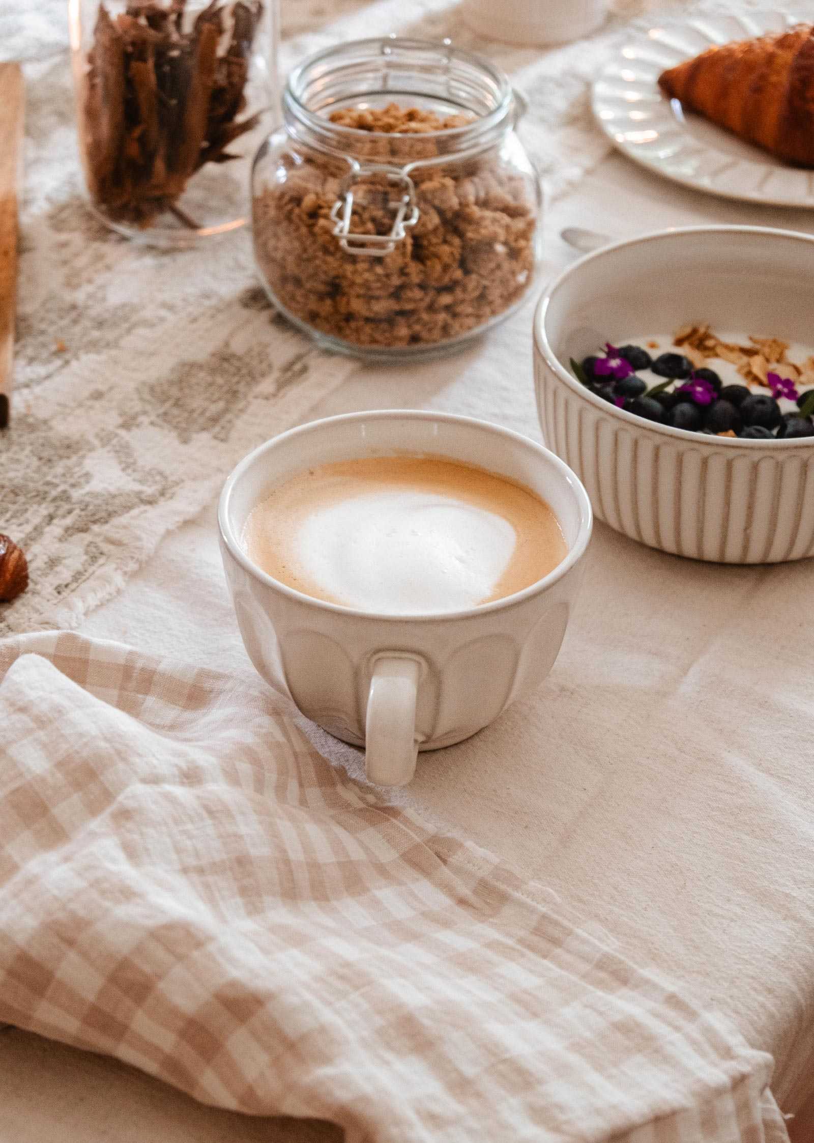 A cup of coffee in the Large Ceramic Latte Mug by Mimi & August sits on a table beside a checked napkin, a bowl of granola and fruit, a croissant on a plate, and a jar of cereal.