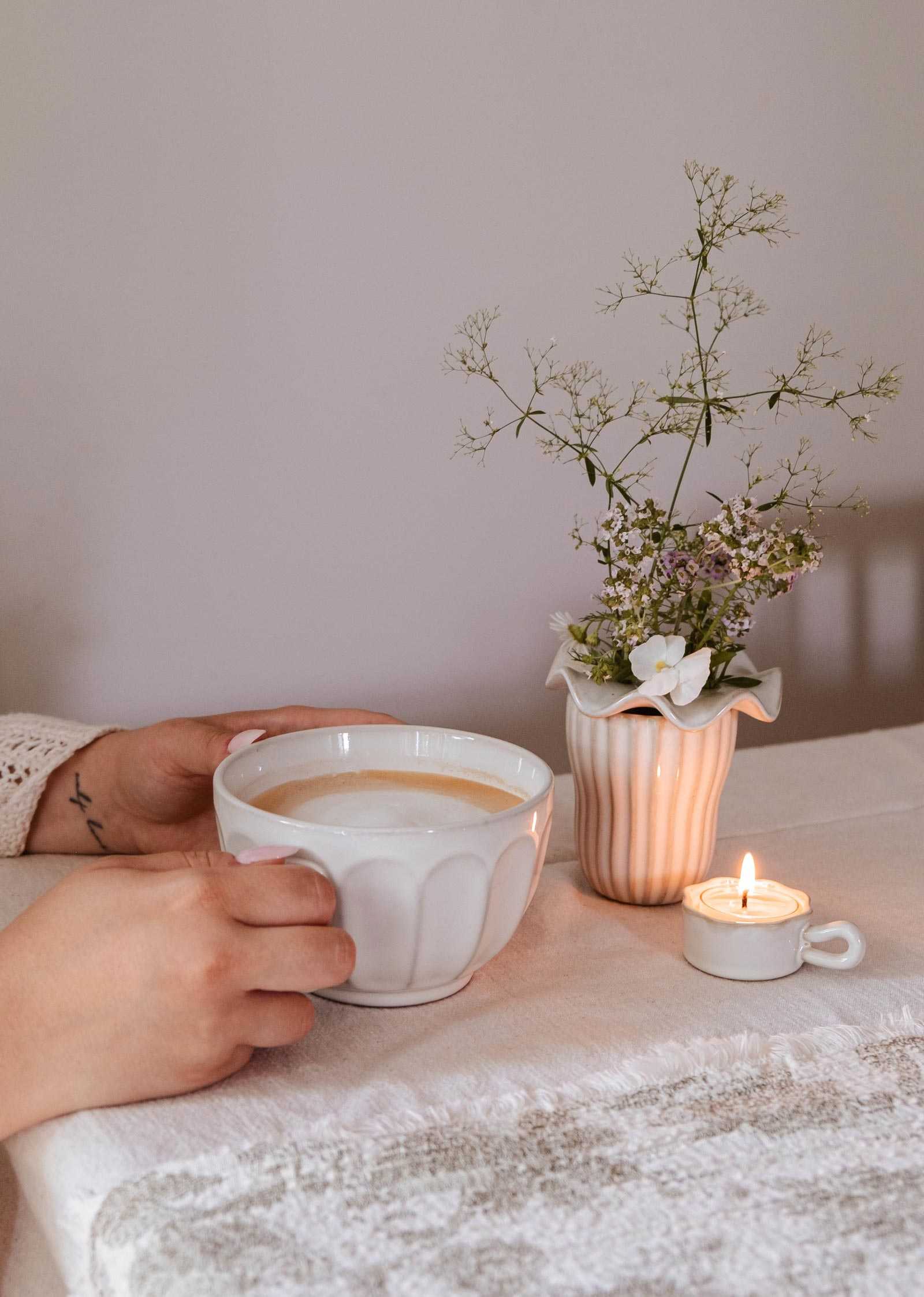 A person holds The Ceramic Latte Mug by Mimi & August filled with coffee on a table beside a small vase of flowers and a lit tealight candle.