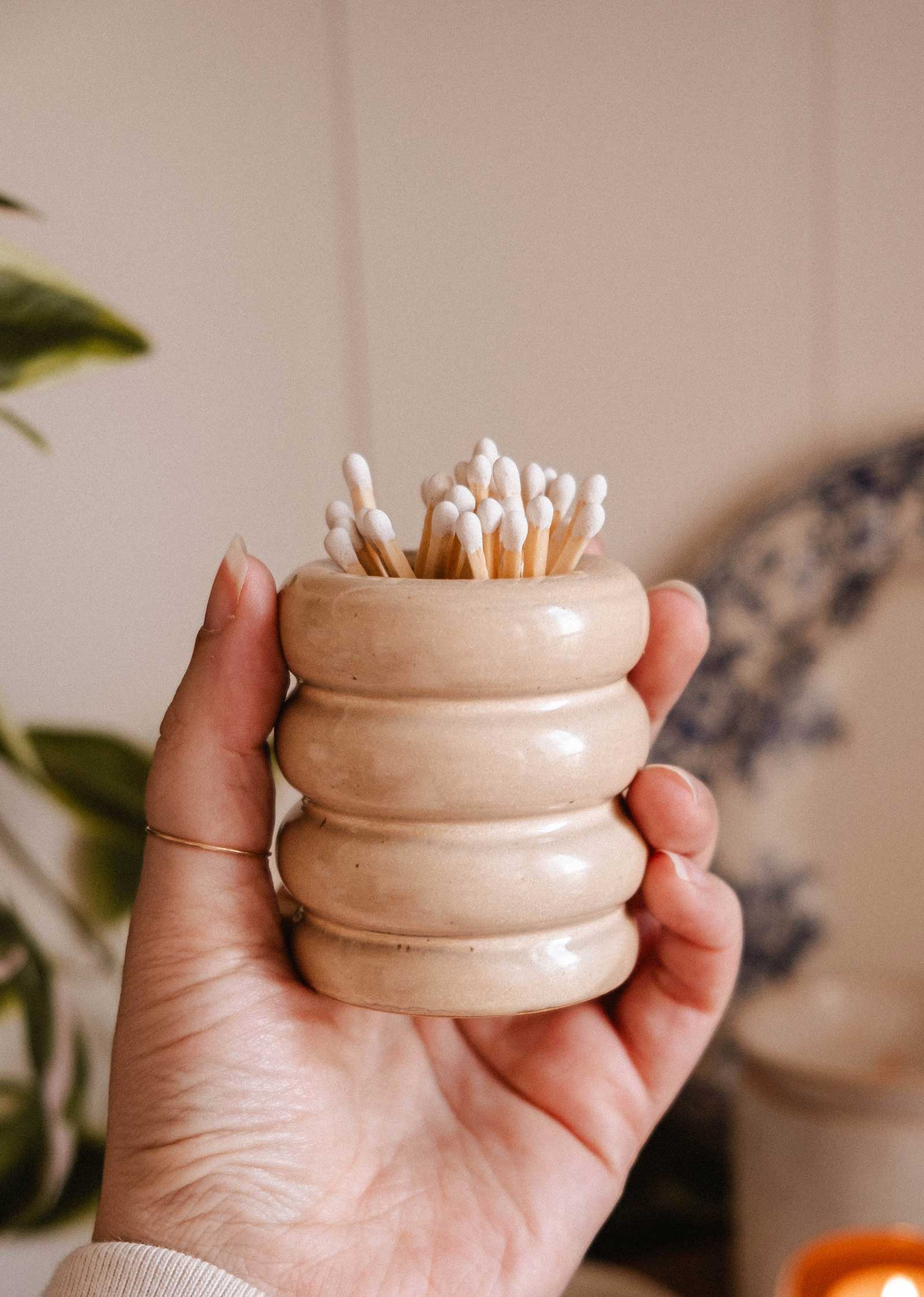 A hand holds the Mimi & August Ceramic Match Pot in beige, featuring a ribbed design and filled with cotton swabs. A blurred plant and plate appear in the background, highlighting the charm of this decorative candle accessory.