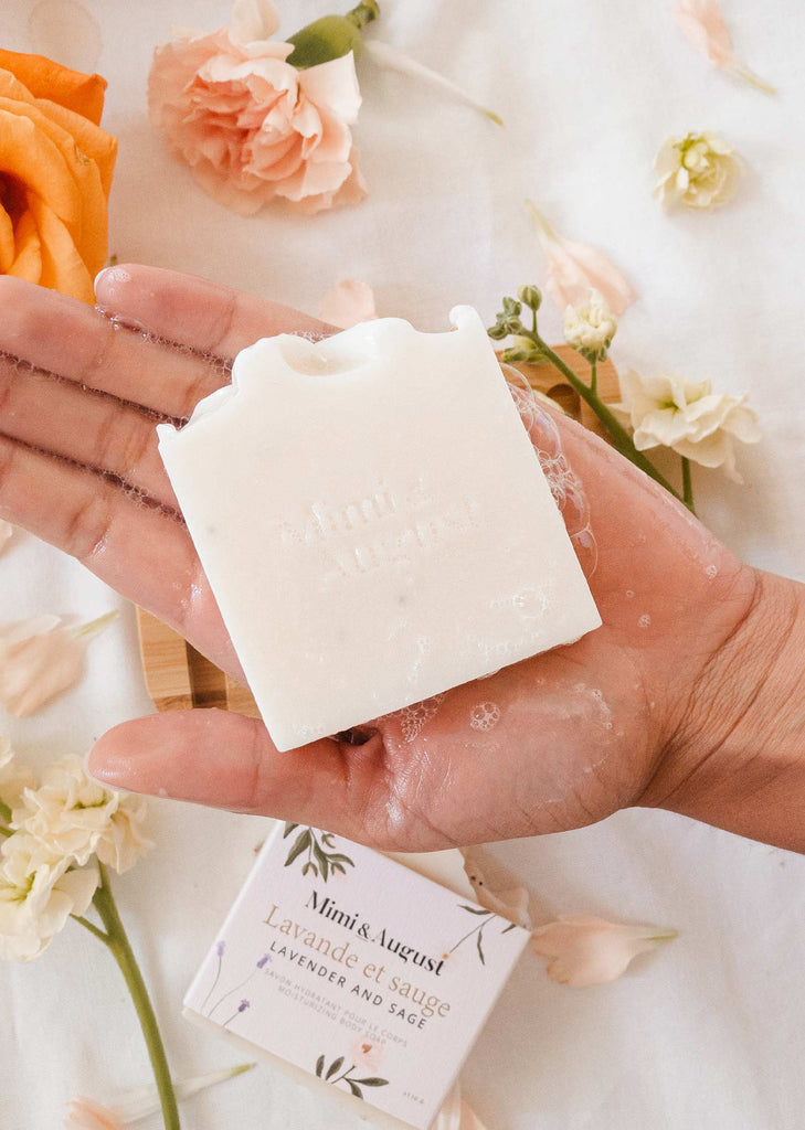A hand holding a Mimi and August Lavender and Sage soap bar next to flowers.