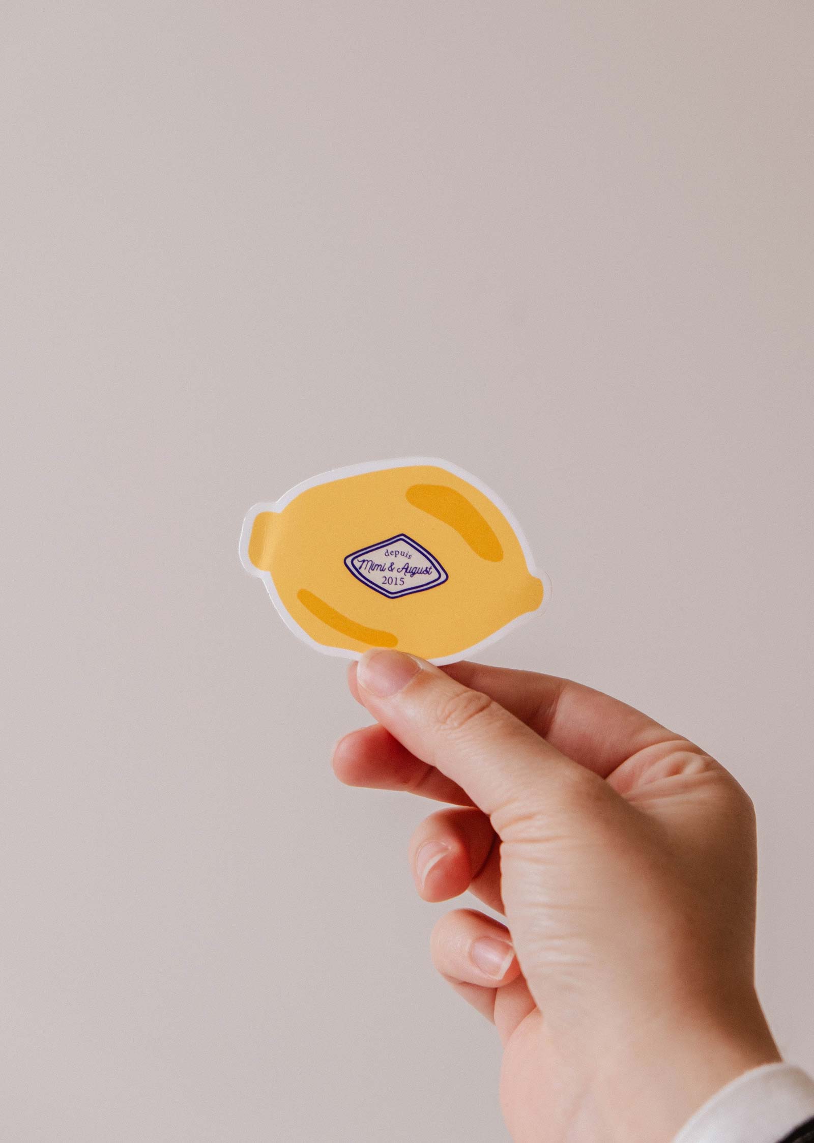 A hand holds the Lemon - Vinyl Sticker by Mimi & August, featuring a vintage-style label in the center, against a plain light background.