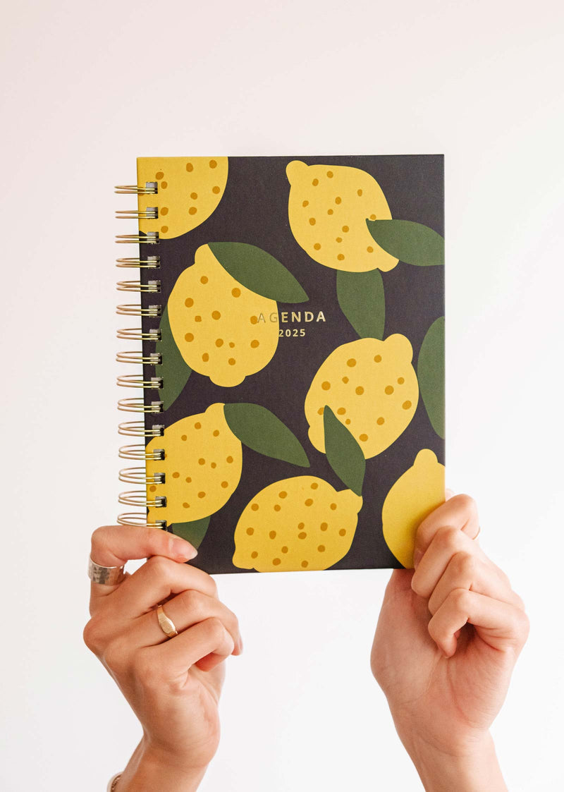 Mains tenant le planning Mimi & August Lemon - Agenda 2025, présentant un motif de citron vibrant avec des feuilles vertes sur un fond noir élégant.