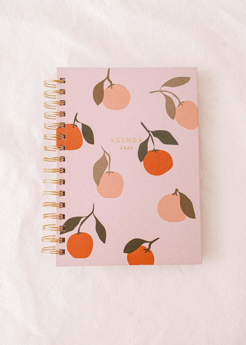 Voici Les Oranges - Agenda 2025 de Mimi & August, un agenda chic doté d'une couverture rose avec un motif dessiné à la main de fruits et de feuilles d'orange, magnifiquement reliée par une spirale dorée.