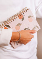 A person in a white sweater holds the Mimi & August Les Oranges - Agenda 2025, featuring a charming hand-drawn orange fruit pattern on its cover.