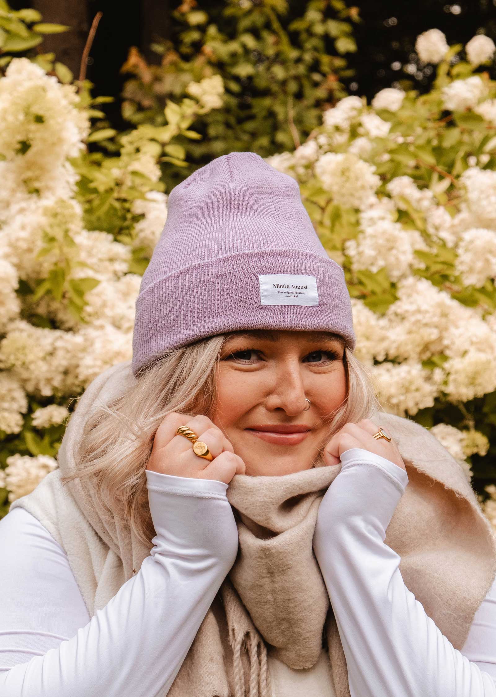 Une personne portant un bonnet lilas Mimi & August et une écharpe beige sourit en tenant son accessoire douillet, debout devant de la verdure luxuriante et des fleurs blanches.